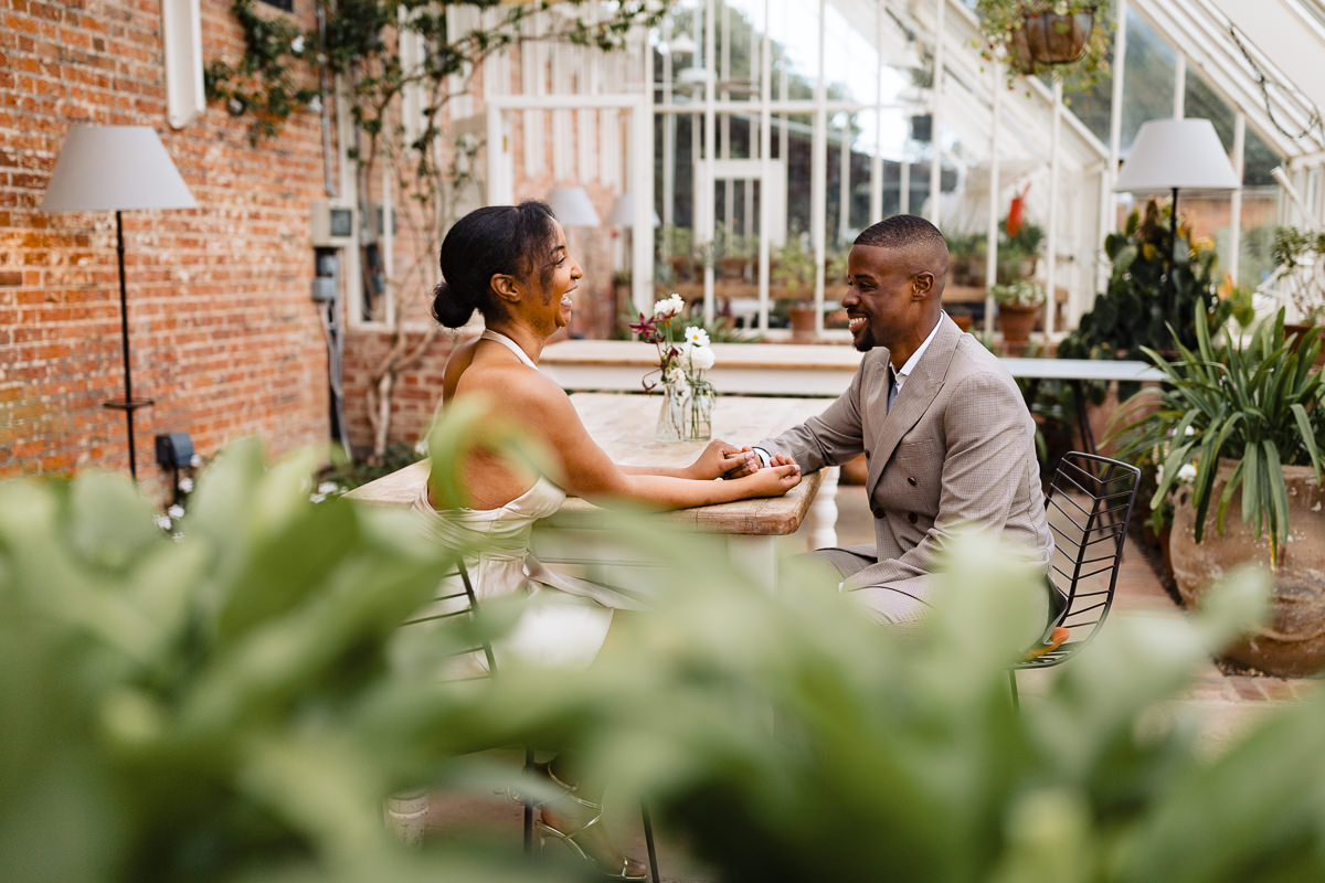 young black hetrosexual couple sit at a table in the glasshouse at heckfield place, holding hands.