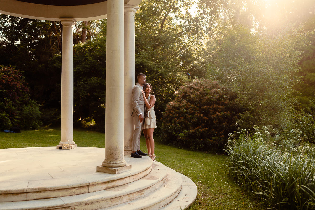 Young heterosexual black couple stand on the top step of the pavillion at Heckfield place with the sun shining on them from above