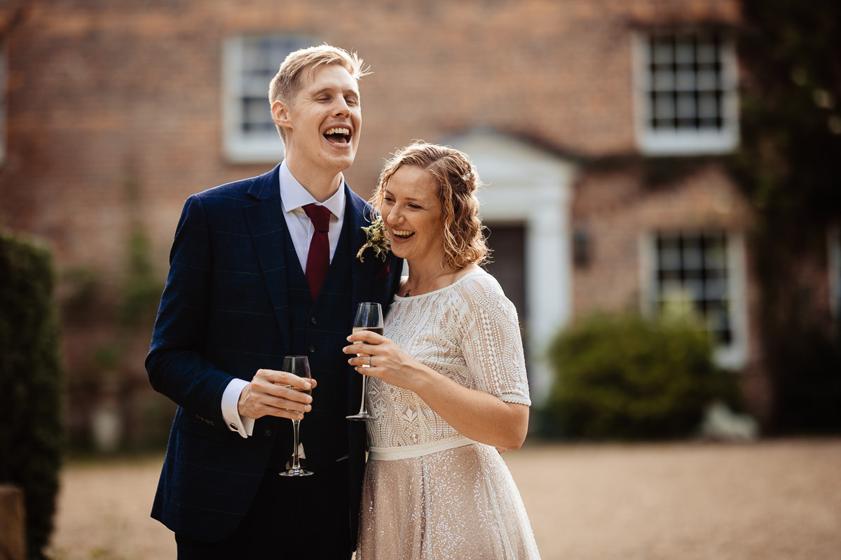 A groom in a navy suit hugs a bride wearing a pink sequinned skirt & lace top in front of Combe manor. They are laughing with relaxed expressions enjoying a moment