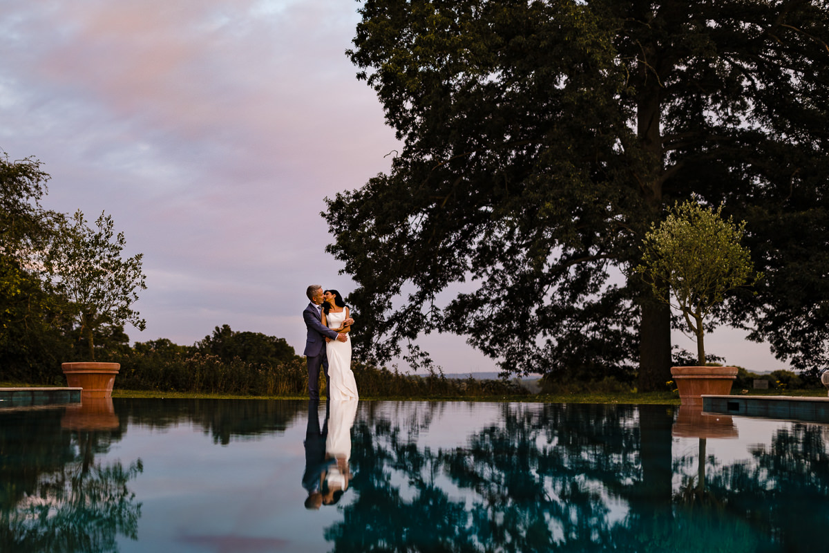 Hetrosexual couple embrace by the swimming pool at Elcot Park with a pink sky behind them