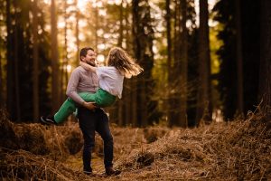 He is smiling holding her at waist height swinging her round in a woodland lit by evening sunshine