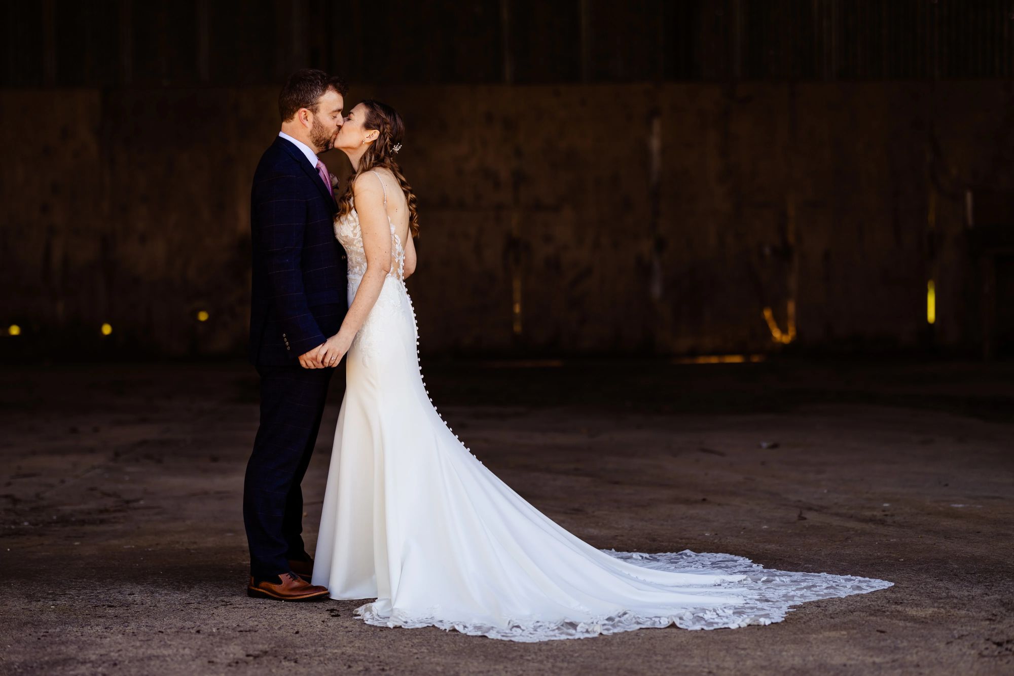 Claire & Edd share a kiss in a dimly lit disused barn at Clock Barn. Claire wears a fitted gown with a long lace train, and Edd is wearing a navy suit, holding hands as they stand face to face.