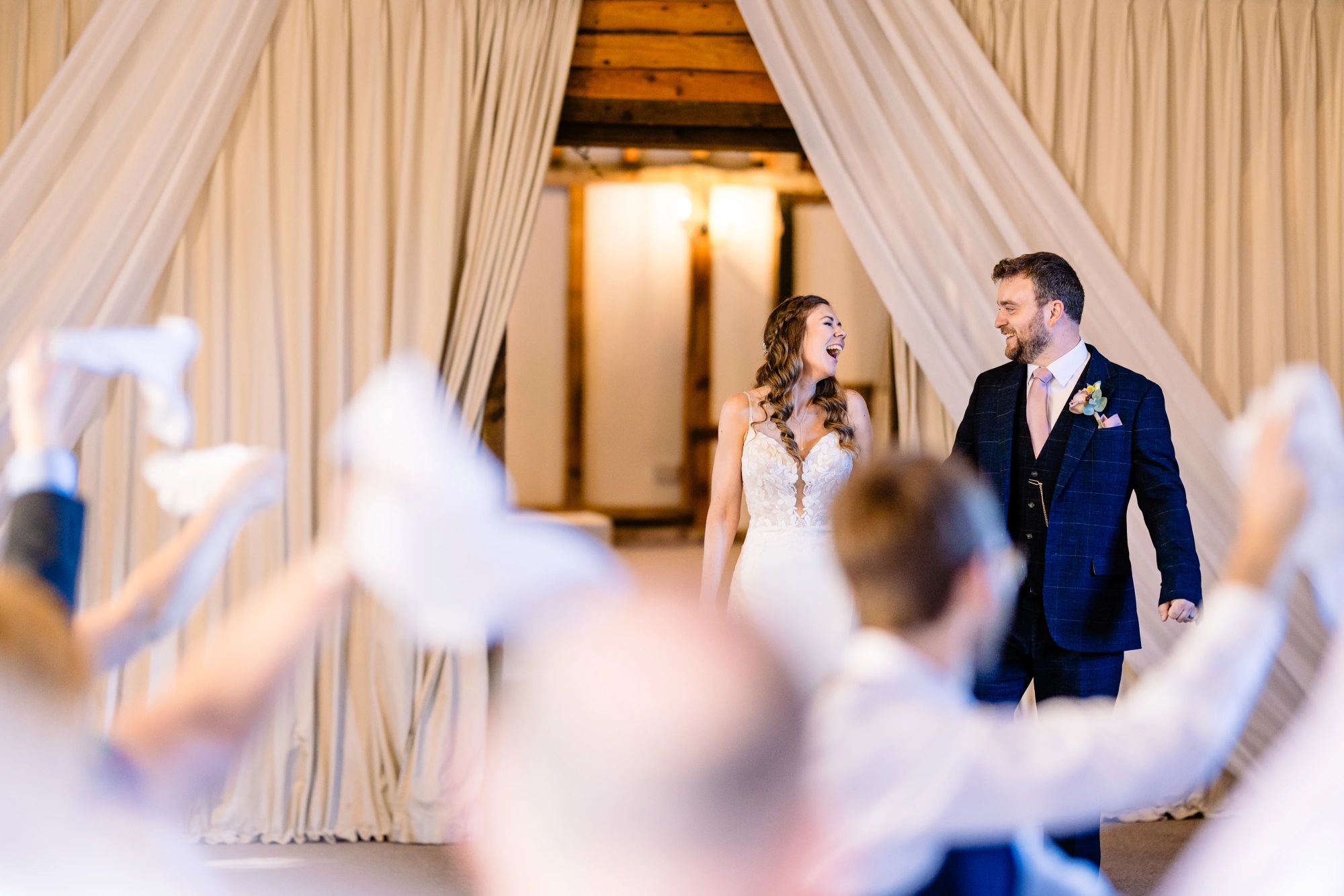 Claire & Edd stand side by side inside the Clock Barn with soft drapery, looking at each other and laughing, as guests wave napkins in the foreground.