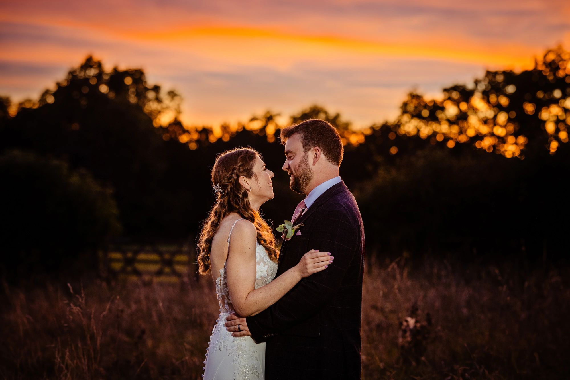 Claire & Edd embrace in the field behind Clock Barn at sunset, framed by a glowing orange sky. They look into each other’s eyes, with soft silhouettes of trees in the distance.