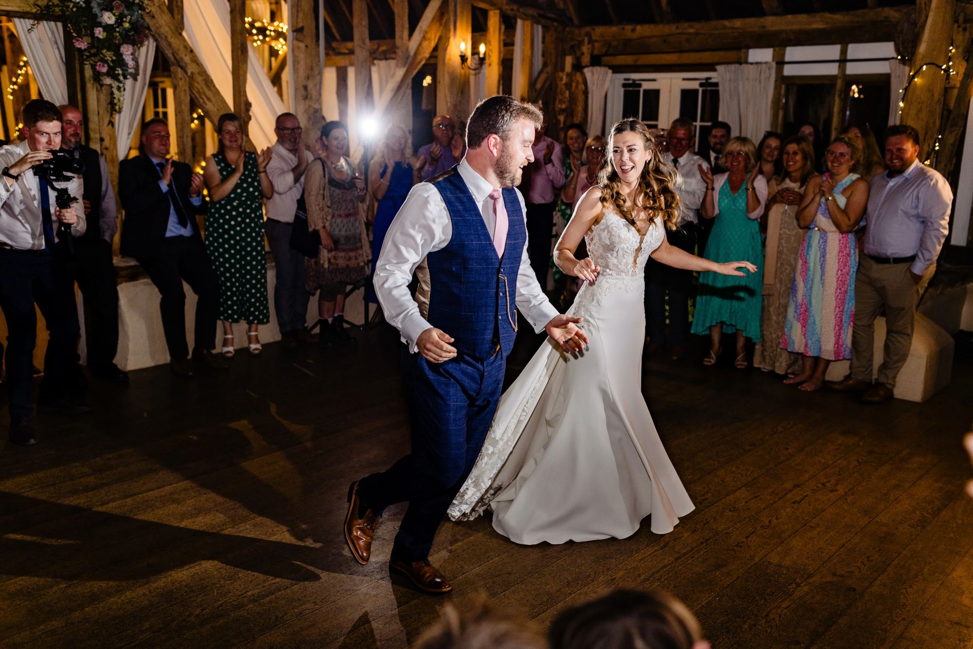 Claire & Edd begin their 1st dance together on a wooden floor, smiling as guests clap and cheer around them.