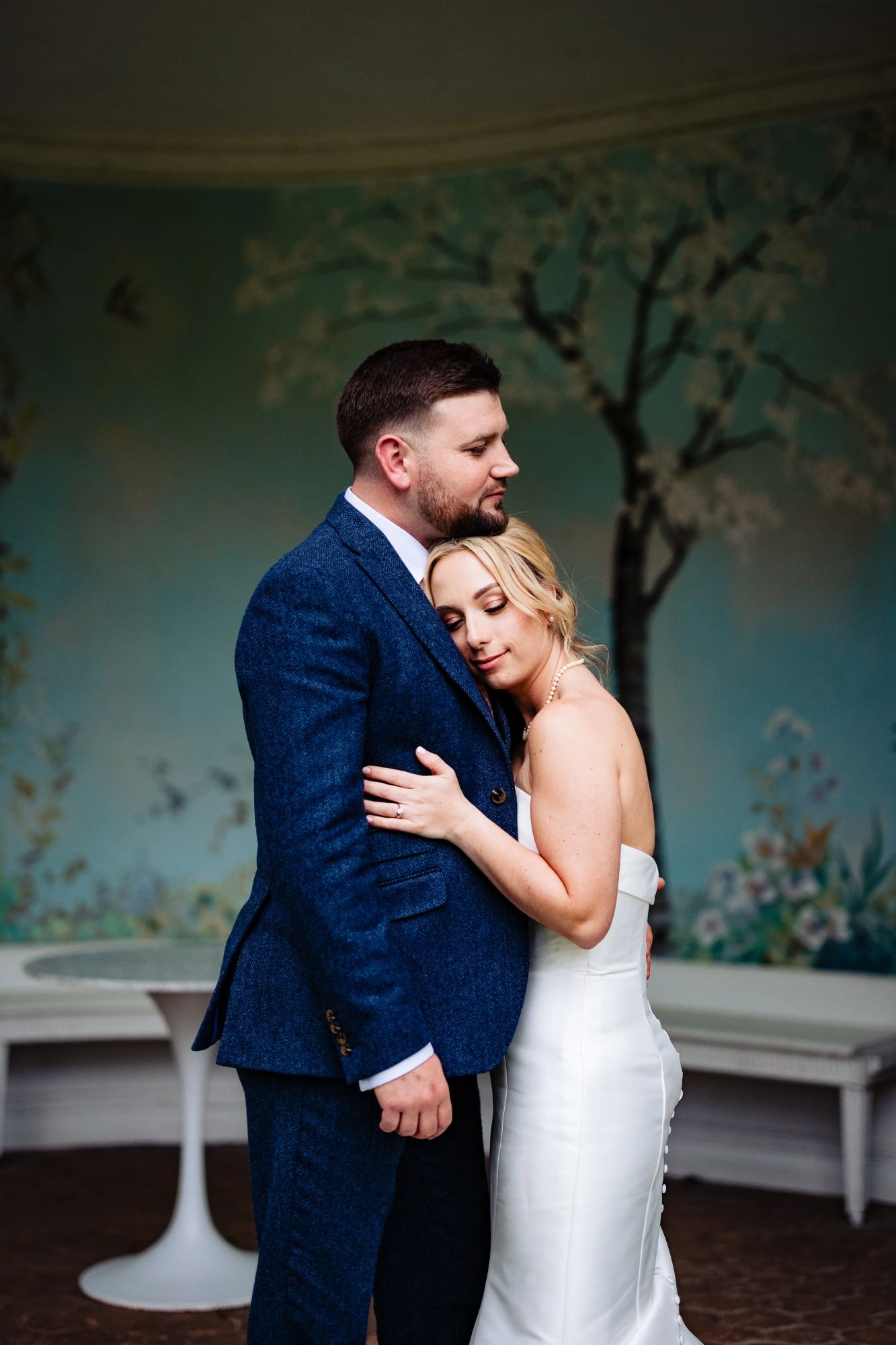 Jamie, in a textured navy suit, embraces Jess who leans her head on his chest, both standing in front of a painted floral mural. Her strapless gown forms a sleek silhouette against his suit.