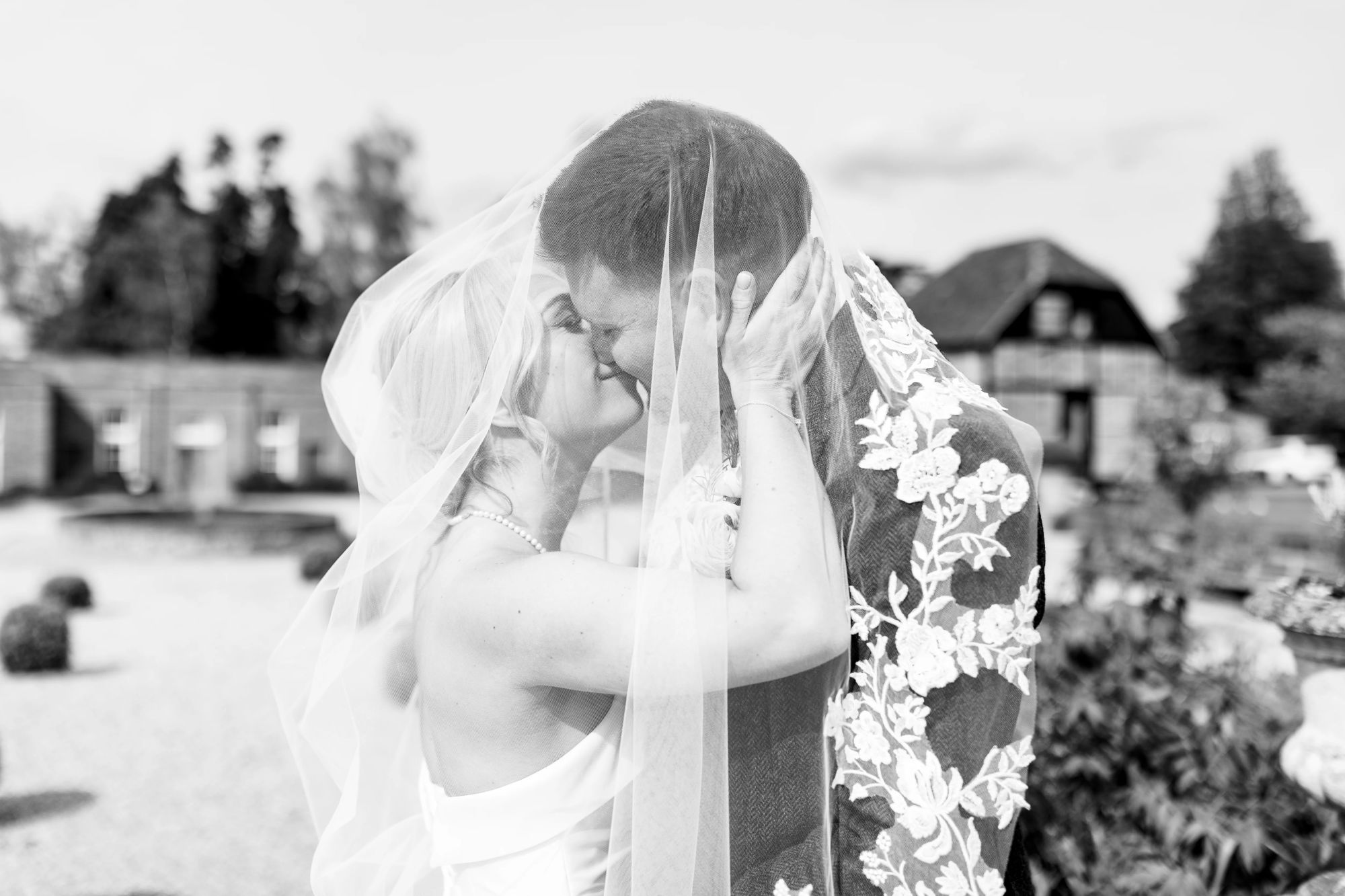 Black-and-white portrait of Jess and Jamie sharing a tender kiss beneath her lace-trimmed veil, with the fountain’s water spray softly blurred in the background.