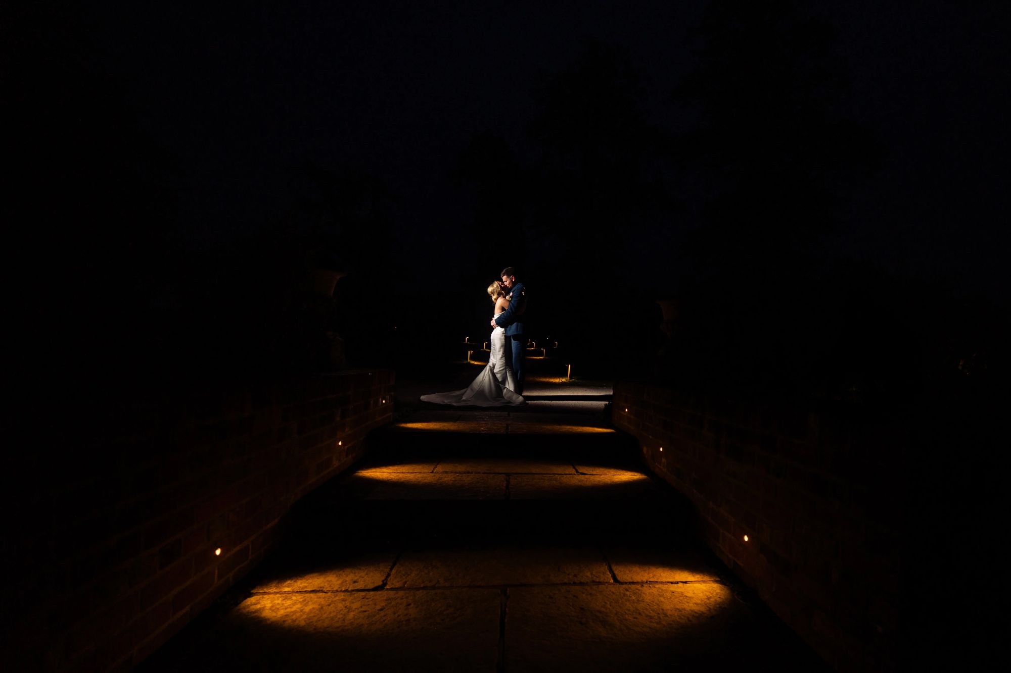 Jess and Jamie standing on an illuminated pathway at Wasing Park after dark, the bride’s dress and the groom’s suit softly lit against the surrounding night by off camera flash.