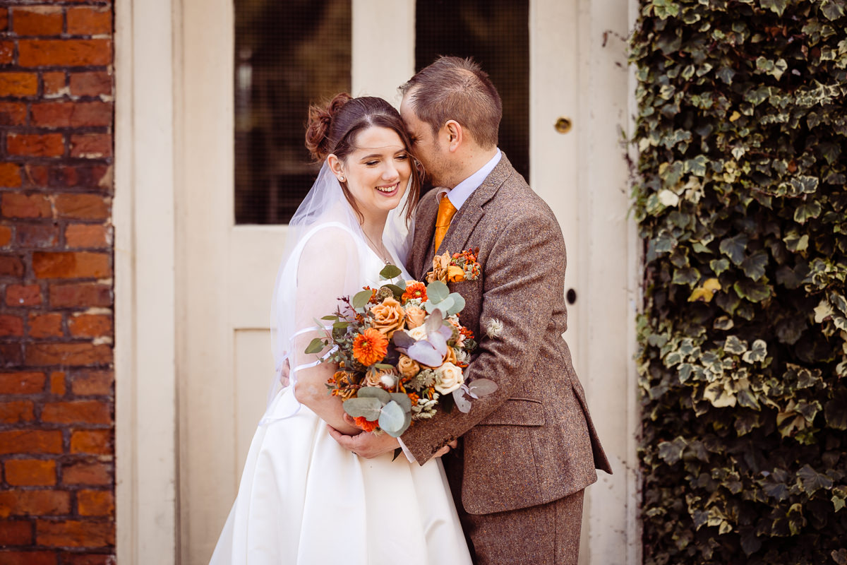 A groom wearing a brown tweed sujit whispers shyly into the ear of his bride who smiles sweetly. They are in front of the clockhouse at Stanlake Vineyard