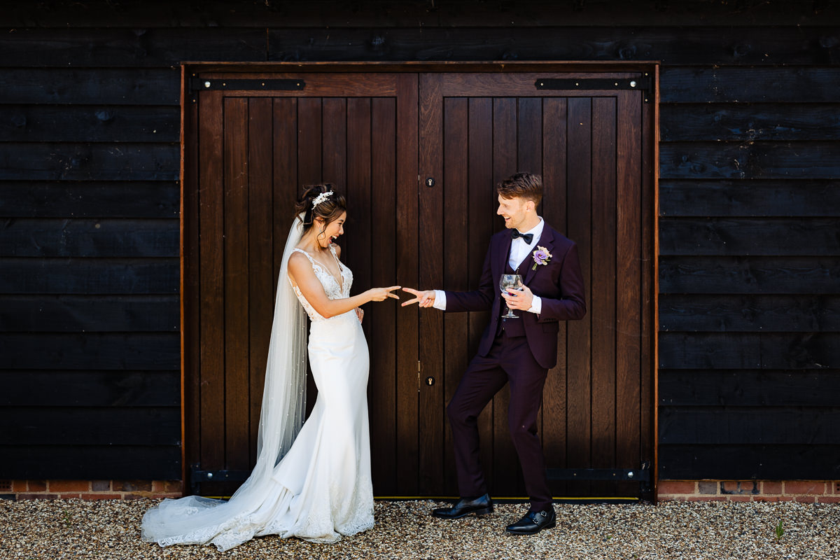 An oriental bride in a fitted white wedding dress & full veil plays rock paper scissors with her groom. He is wearing a purple suit & they are in front of wooden doors at Lillibrooke manor. They're laughing playfully